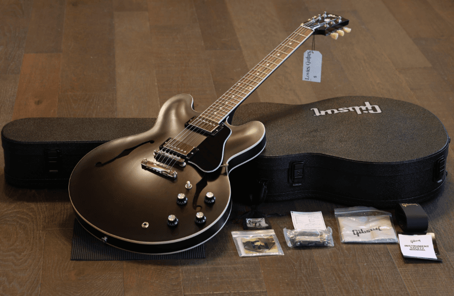 An unplayed 2025 Gibson ES-335 in Satin Smokehouse lying beside its open hard shell case, with the case candy and safety manual laid out on a wood floor